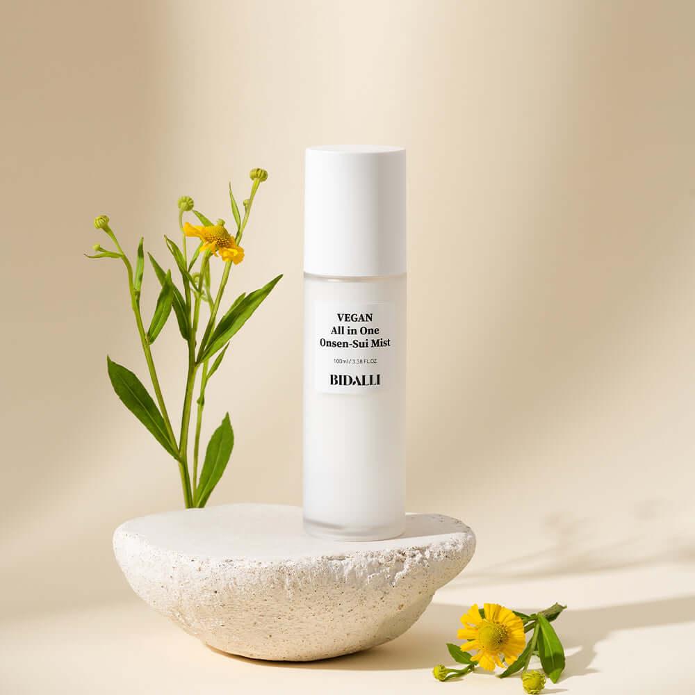 Bidalli Vegan All in One Onsen-Sui Mist placed on natural stone with yellow flowers, hydrating vegan facial mist inspired by onsen water and botanical skincare