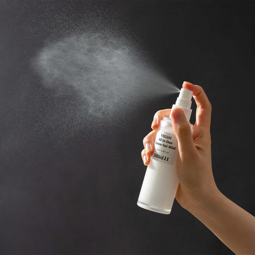 Hand spraying Bidalli Vegan All in One Onsen-Sui Mist in fine mist cloud against black background, demonstrating ultra-fine hydrating facial mist for refreshed and dewy skin