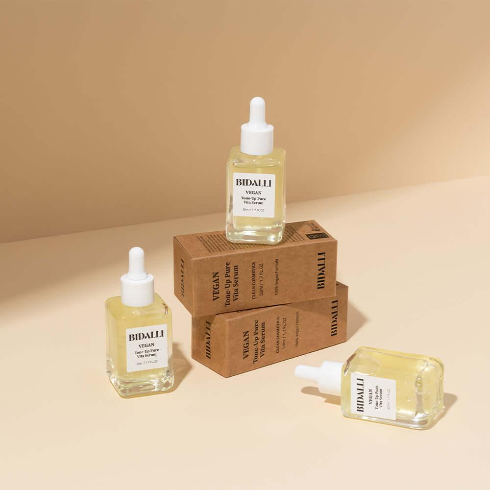 Four Bidalli Vegan Tone-Up Pure Vita Serums displayed with kraft paper boxes on beige background, clean and minimal skincare product scene