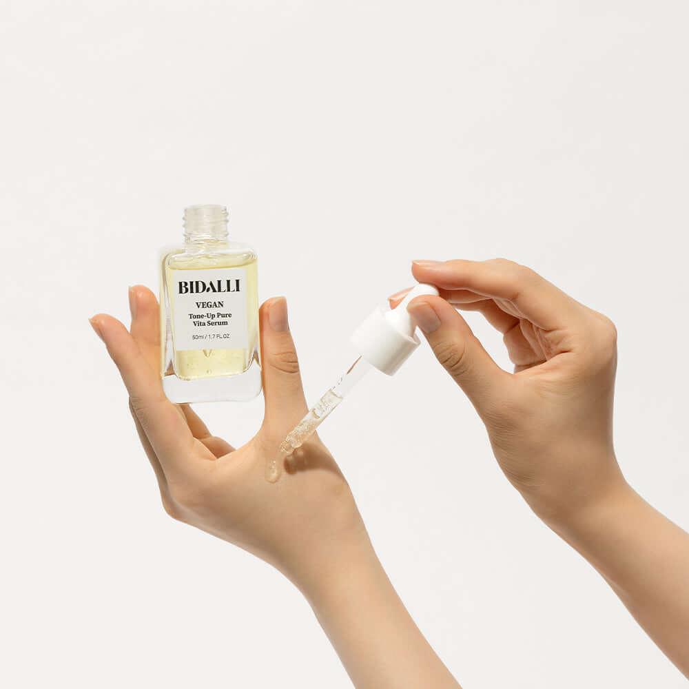 Hand applying Bidalli Vegan Tone-Up Pure Vita Serum with dropper on wrist, minimal skincare product demonstration on white background